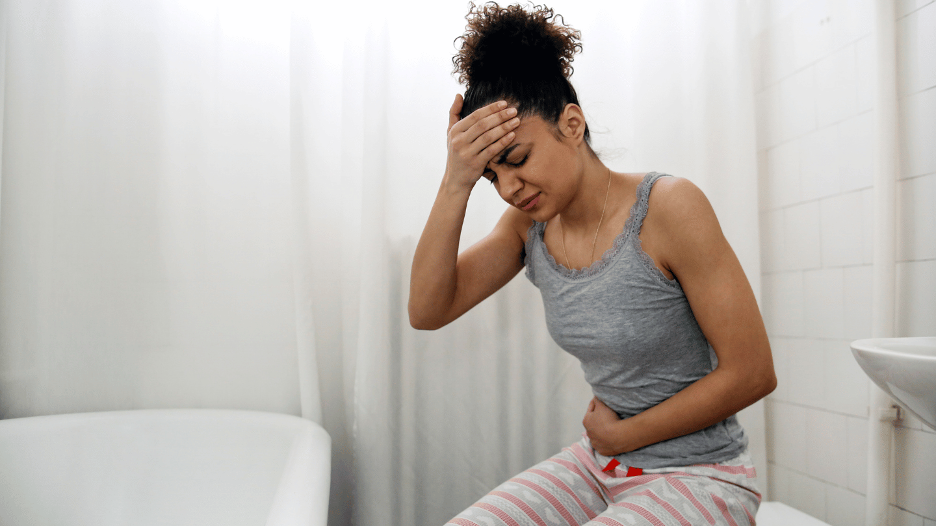 Woman experiencing abdominal pain and constipation while sitting in a bathroom, holding her stomach and head in discomfort.