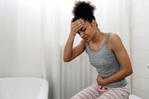 Woman experiencing abdominal pain and constipation while sitting in a bathroom, holding her stomach and head in discomfort.